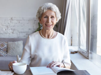 Horizontal shot of cheerful happy retired European gray haired lady relaxing indoors with cup of fresh hot coffee and interesting book. Charming female pensioner reading article in fashion magazine